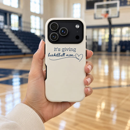 Hand holding a cream iPhone case with navy ‘it’s giving basketball mom.’ script and a heart flourish, in a basketball gym.