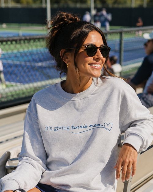 A woman sitting on outdoor stadium bleachers at a tennis court, wearing a light gray sweatshirt with the text 'it's giving tennis mom' and a heart in blue script. She is also wearing navy athletic shorts and sunglasses, holding a white water bottle.