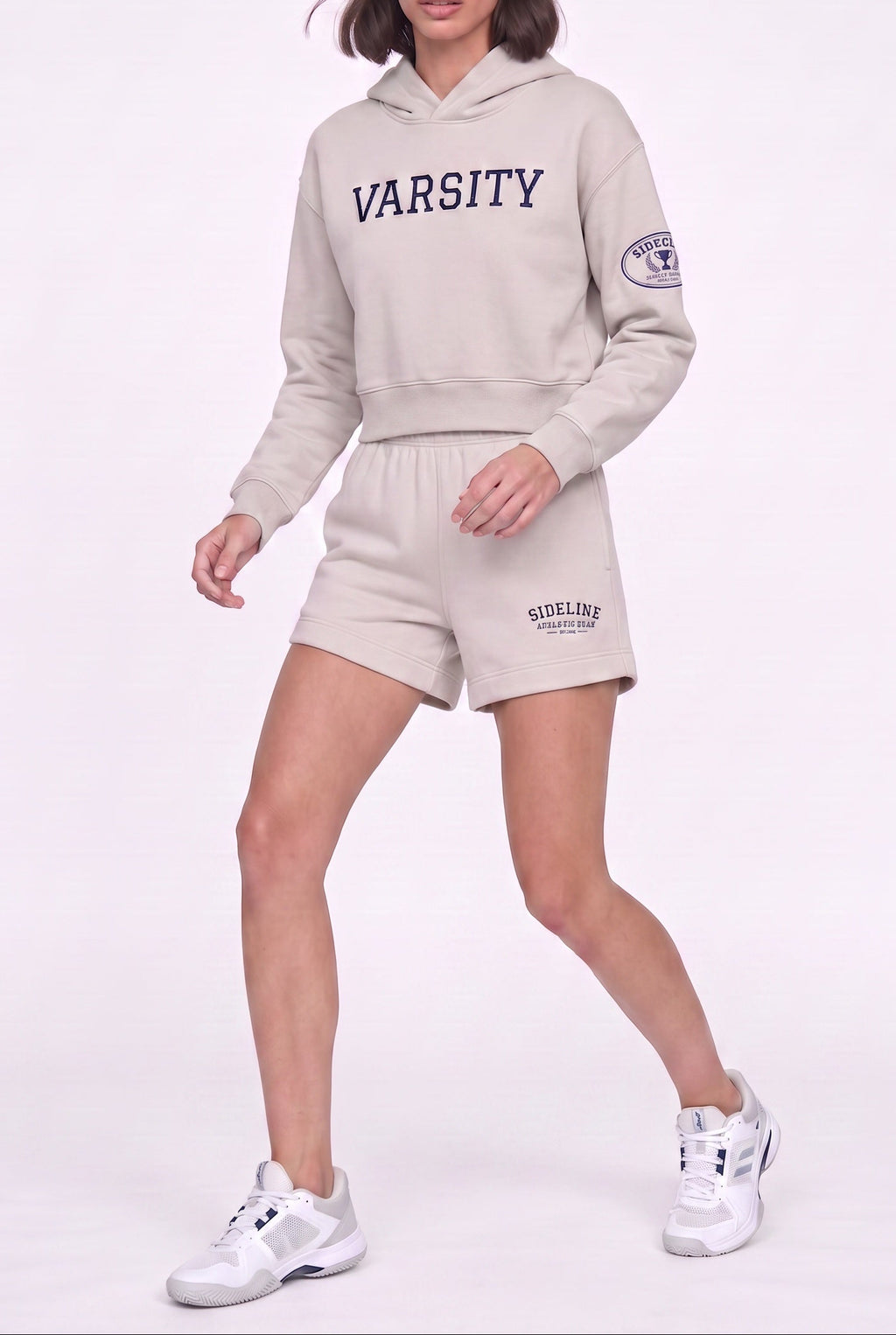 A model wearing a light beige athletic tracksuit featuring a cropped hoodie with 'VARSITY' printed in navy blue on the chest, matching shorts with a 'SHELLEY' logo, and white sneakers, set against a plain white studio background.