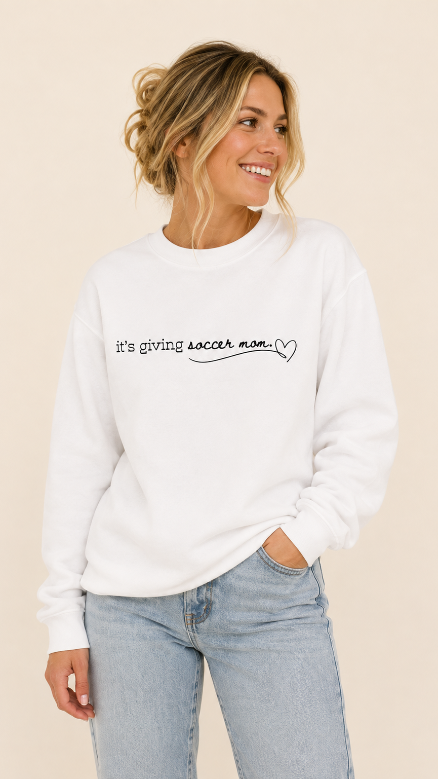 A woman wearing a white crewneck sweatshirt with the black text 'it's giving soccer mom.