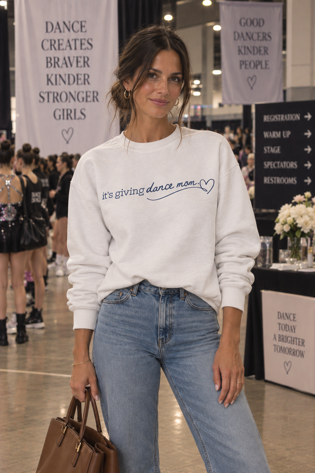 A woman stands in a dance competition or event space wearing a white crewneck sweatshirt with 'it's giving dance mom' printed in blue script, paired with light-wash blue jeans and holding a brown leather handbag.