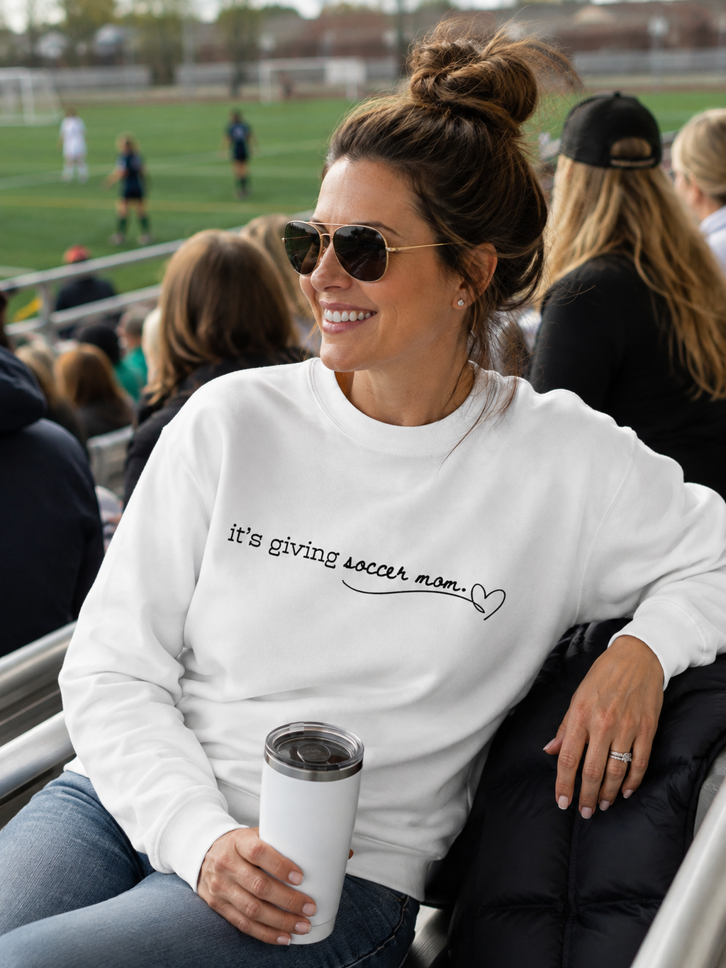 A woman wearing a white crewneck sweatshirt with the text 'it's giving soccer mom.