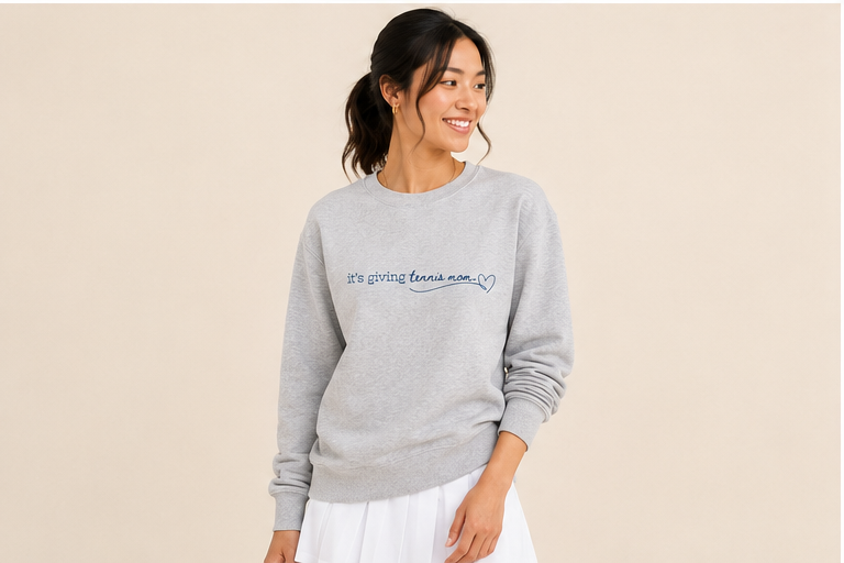 A woman wearing a light gray crewneck sweatshirt with 'it's giving tennis mom.'