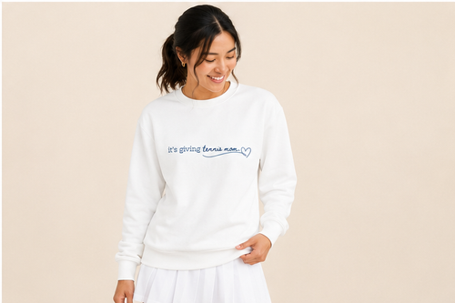 A woman wearing a white crewneck sweatshirt with 'it's giving tennis mom.