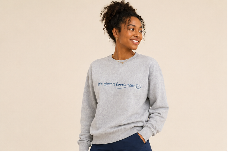 A woman wearing a light gray heather crewneck sweatshirt with blue text reading 'it's giving tennis mom.