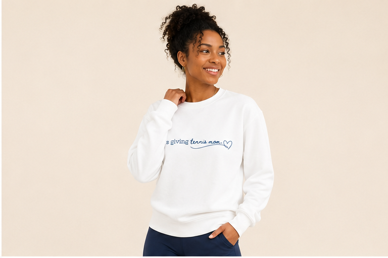 A woman modeling a white crewneck sweatshirt with a blue graphic that reads 'it's giving tennis mom' and a small heart, set against a solid beige background.